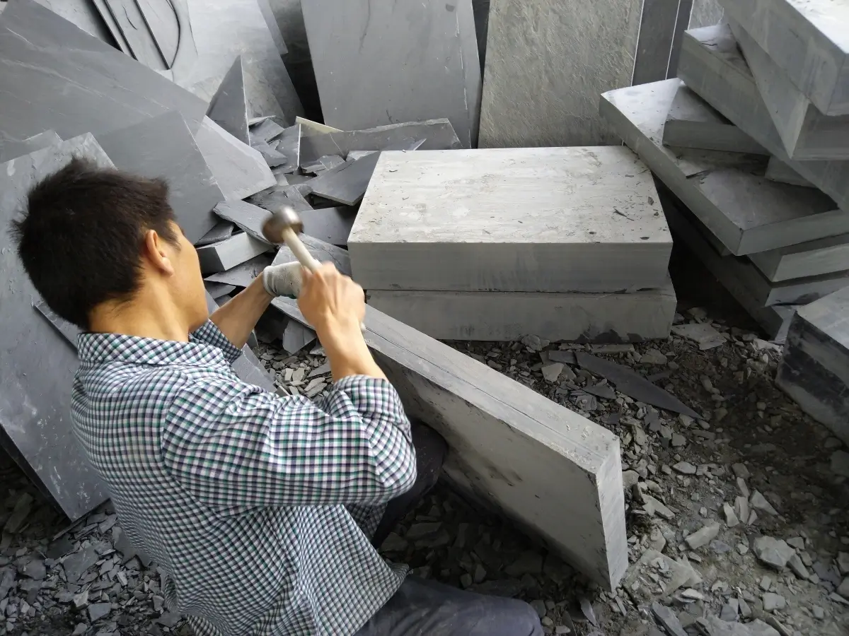 Worker's hands splitting a block of dark grey slate with a chisel and hammer along the natural cleavage plane