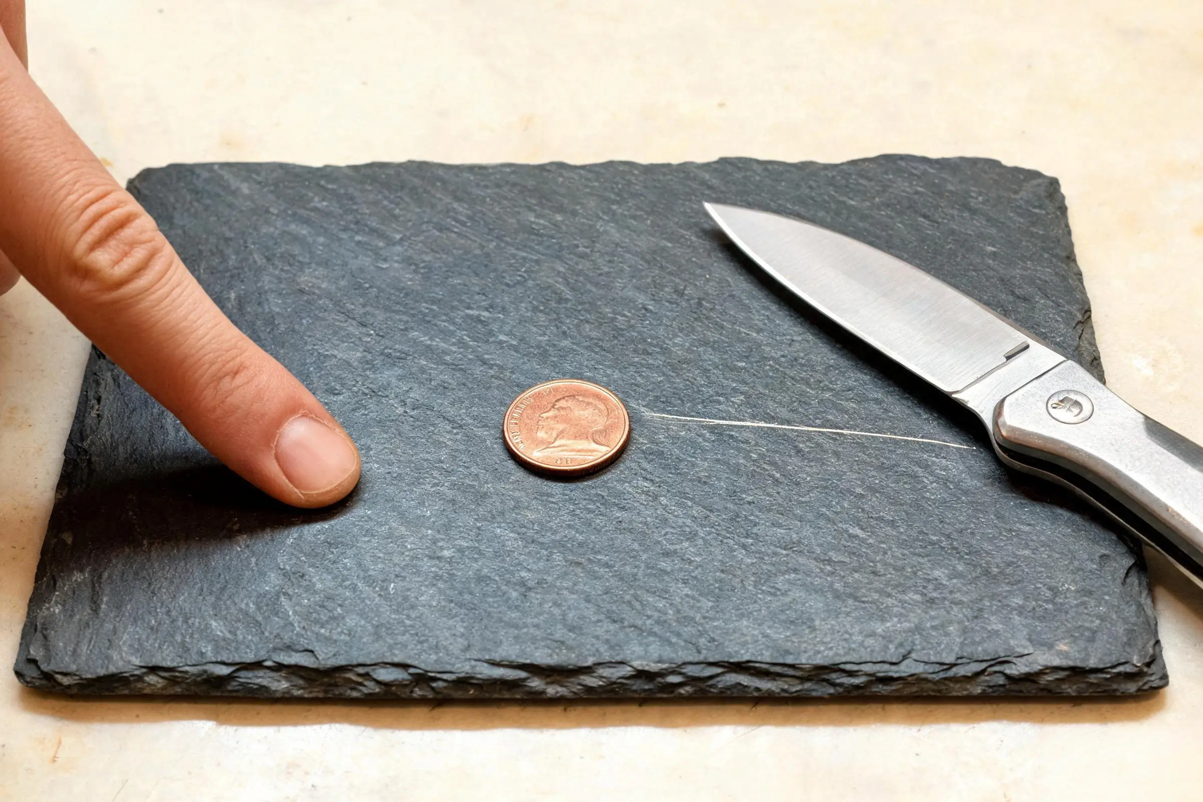 Jiujiang Slate Hardness 1 Three scratch test tools on a dark slate tile: a fingernail, a copper coin and a steel knife blade, showing the Mohs 3-4 hardness range