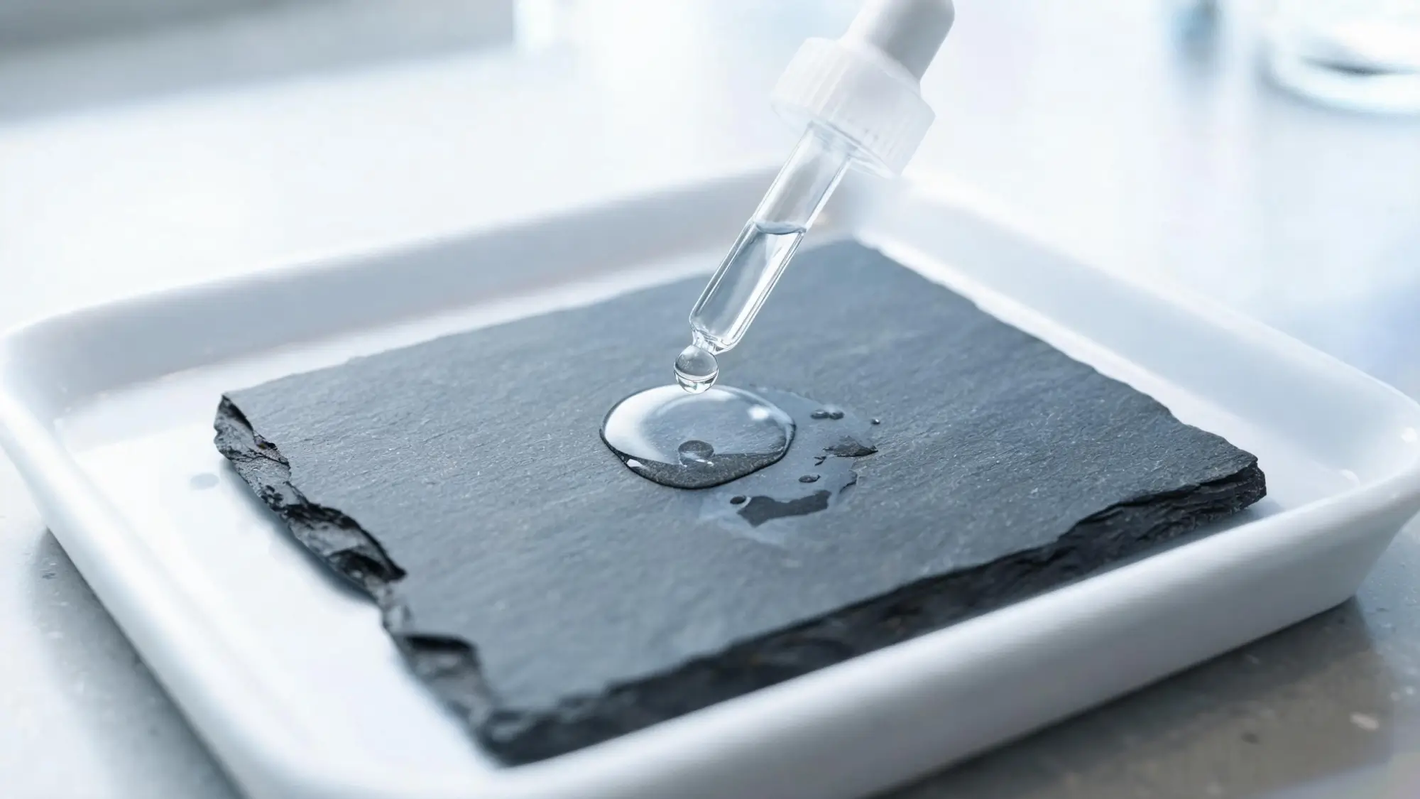 Slate tile in a white laboratory tray receiving a single droplet from a glass dropper during an acid sensitivity test