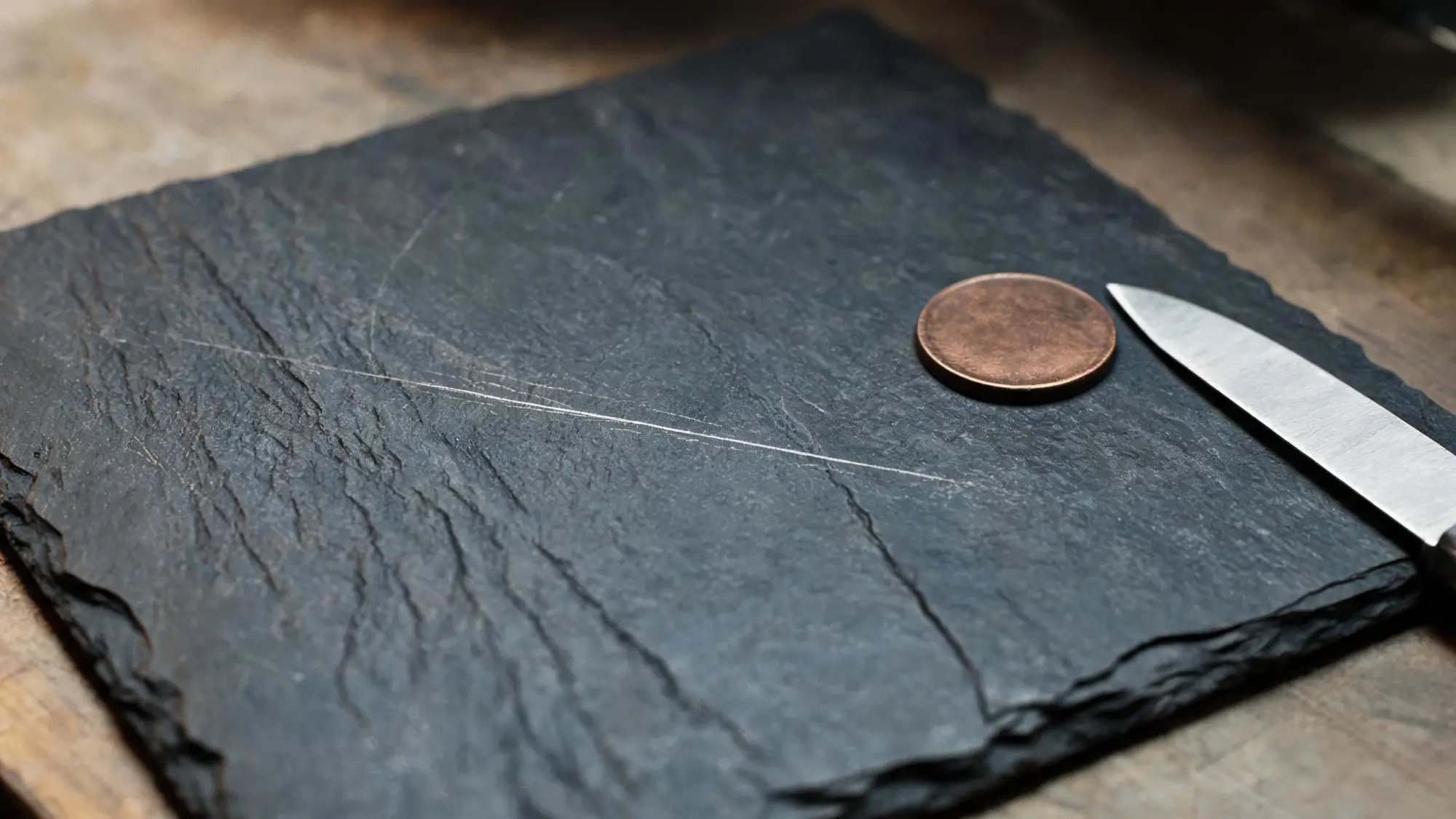 Scratch hardness test on a dark slate tile with a copper coin and steel blade as comparison tools