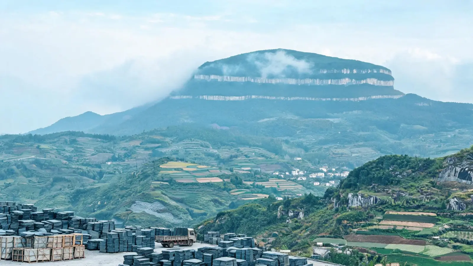 Lushan, Xingzi and the Slate Belt of Jiangxi 1 Panoramic view of the Lushan slate belt — Mount Lushan rising in cloud beyond green foothills, farmland and villages, with a slate yard of stacked blocks and crates in the foreground, Jiangxi China
