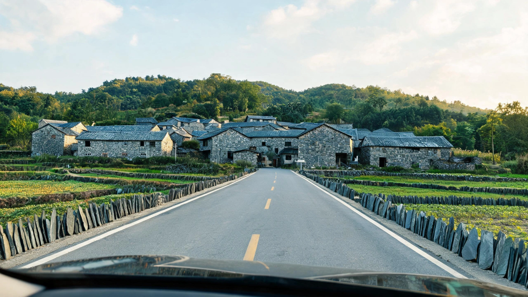 Jiujiang Slate Map 5 A rural road through the Jiujiang slate region approaching a village with grey-blue slate walls, slate roofs and stone-lined field boundaries, green hills behind, in Jiangxi China