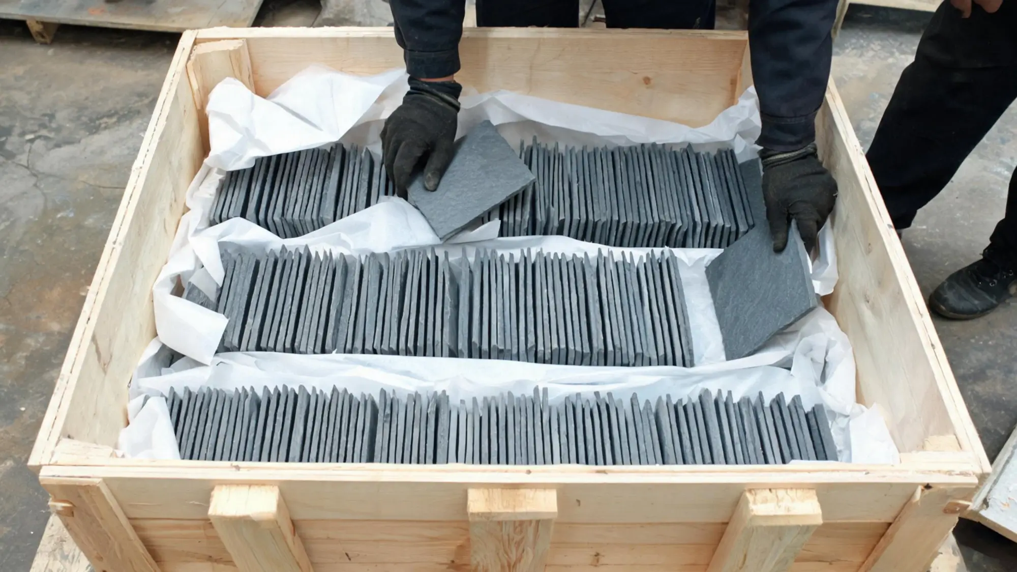 Workers packing grey roofing slate tiles standing upright into a wooden export crate lined with protective paper