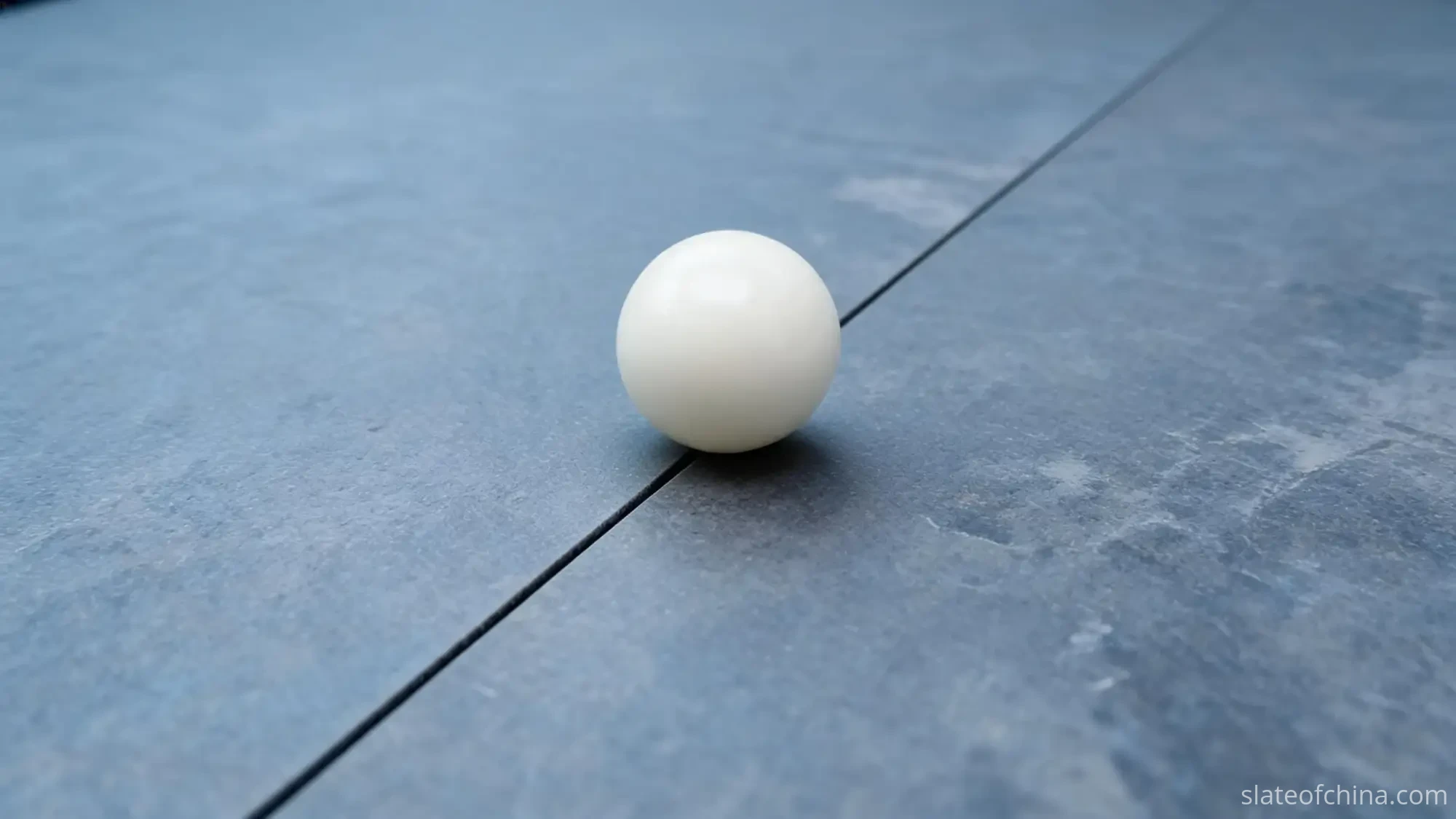 Single white billiard ball resting on honed blue-grey slate with its contact point on the hairline seam between two panels