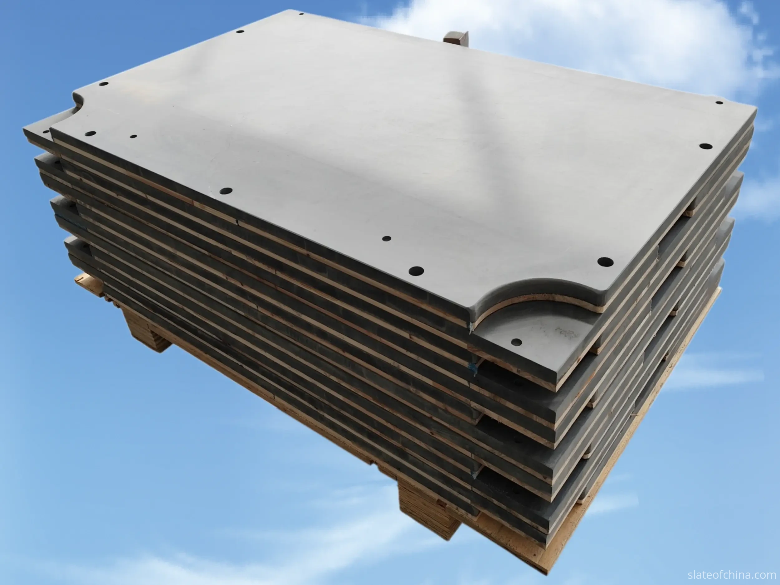 Stack of finished slate panels on a pallet with protective spacers, the top panel showing countersunk mounting holes and shaped cutouts along its edge