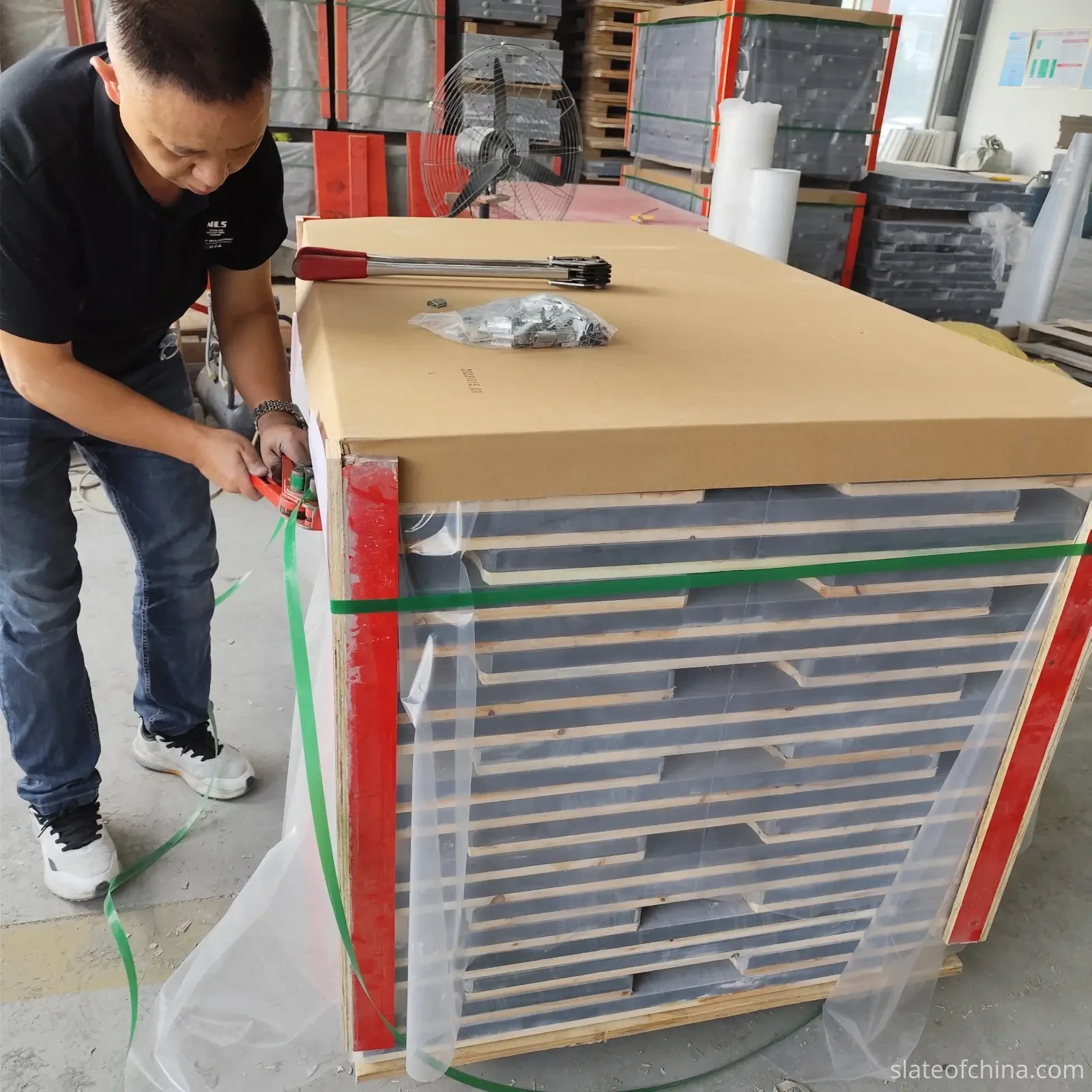 Export Wooden Crate Packing 1 Worker applying green plastic strapping to a palletized stack of billiard slate panels laid flat, with red corner guards, wooden lath separators between layers, and kraft cardboard top cap