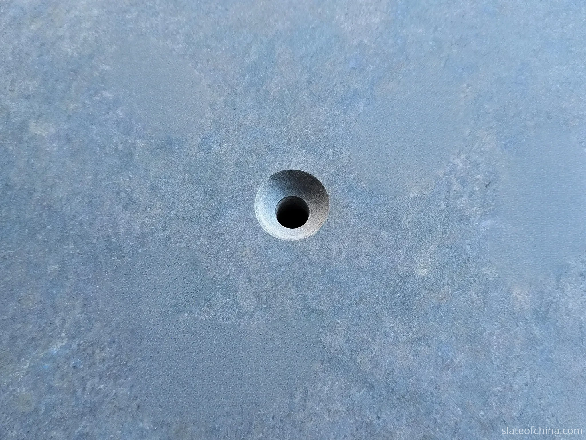 Top-down close-up of a precisely machined countersunk hole in a blue-grey billiard slate surface showing the conical recess