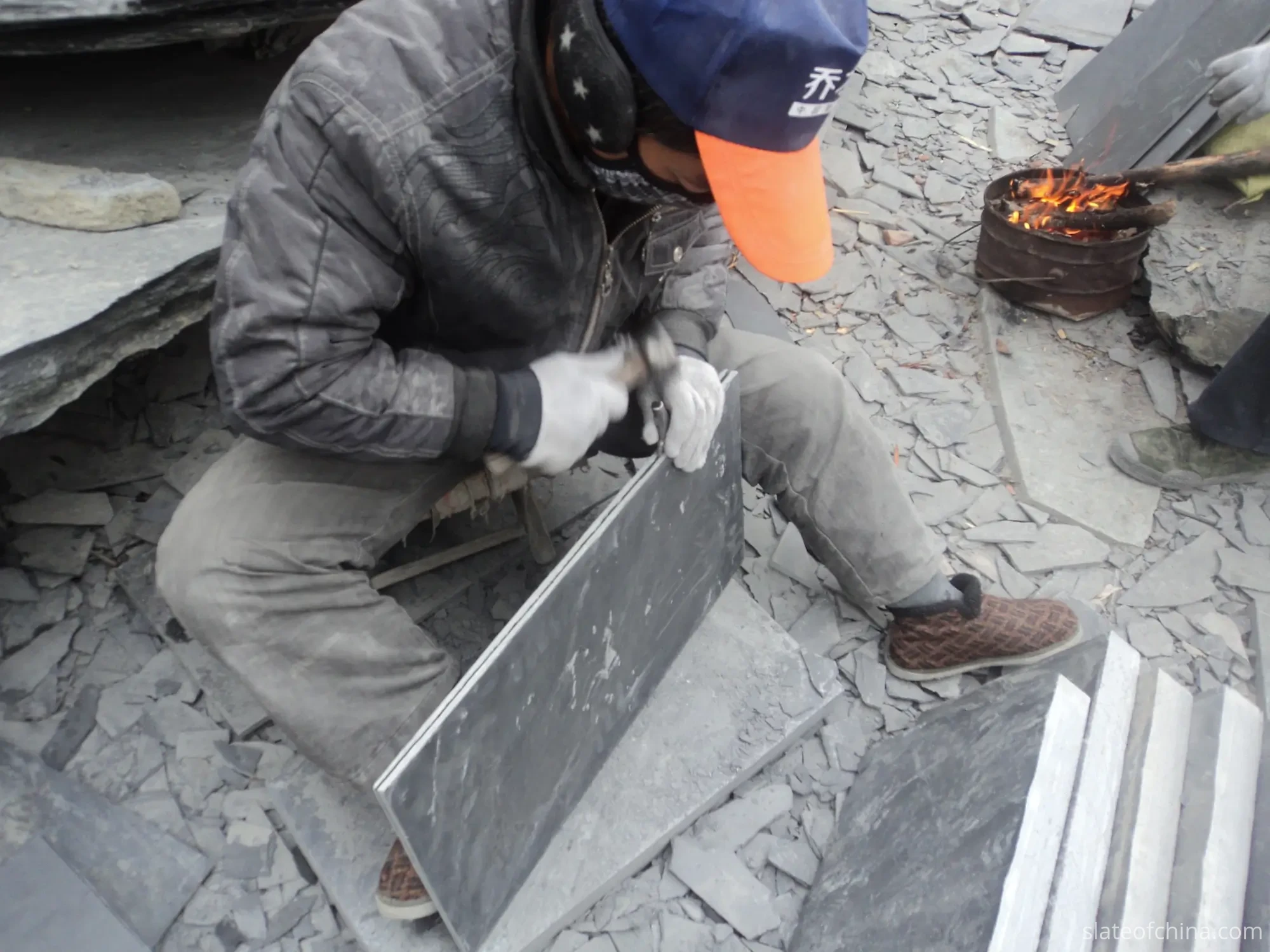 Two older craftsmen hand-splitting slate with hammer and chisel in an open family workshop yard, thin split tiles stacked by their feet