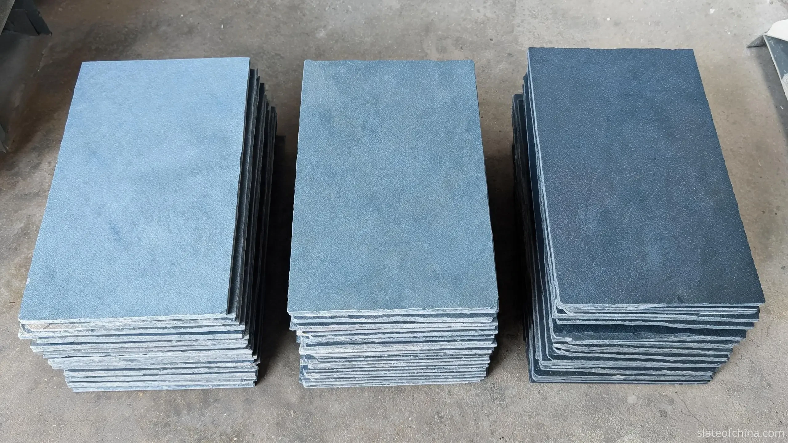 Three stacks of blue-grey roofing slate sorted by tone into light, medium and darker groups on a workshop floor