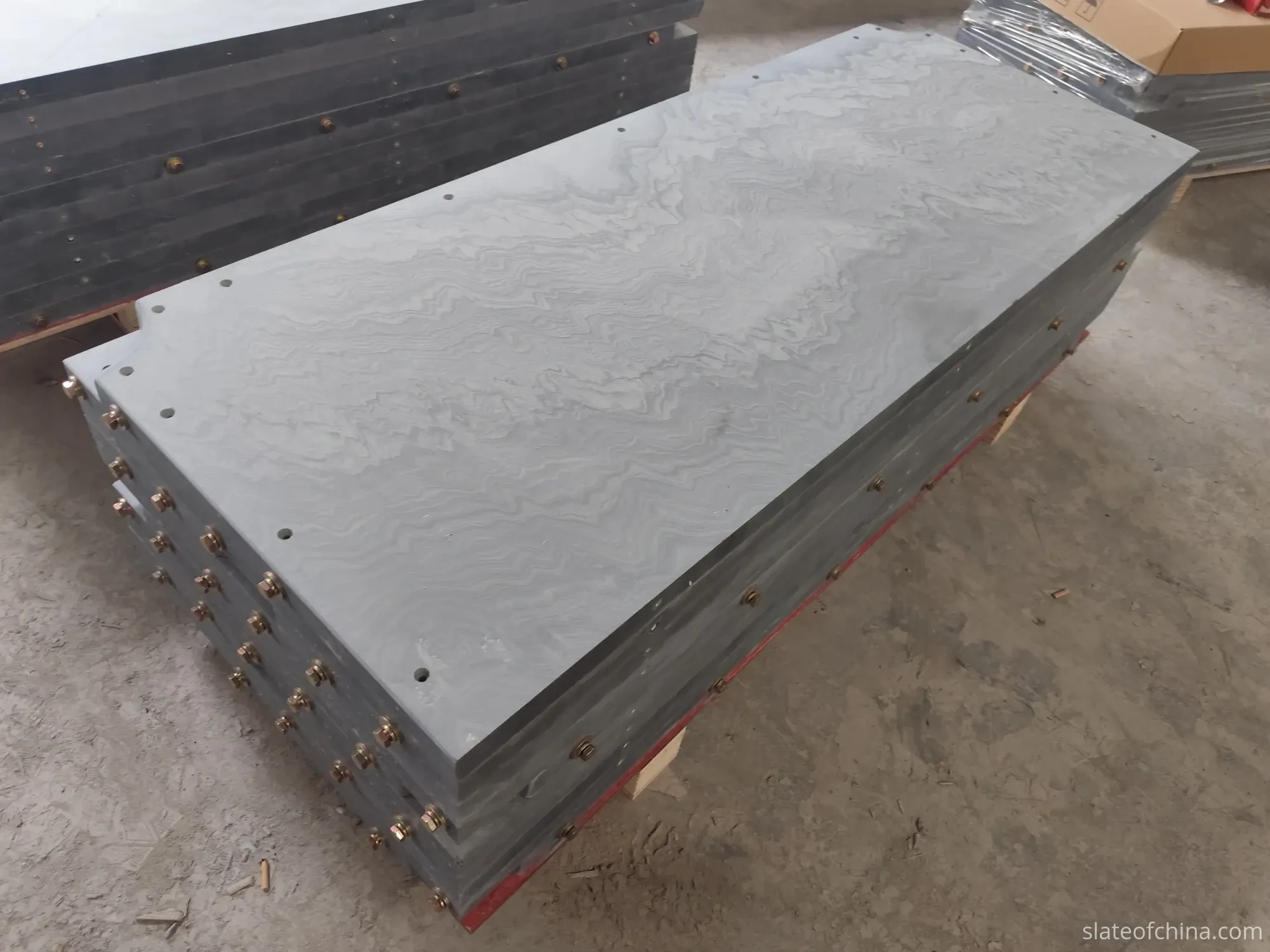 Finished billiard table slate panels stacked on timber support blocks in an OEM workshop, machined edges and bolted edge hardware visible