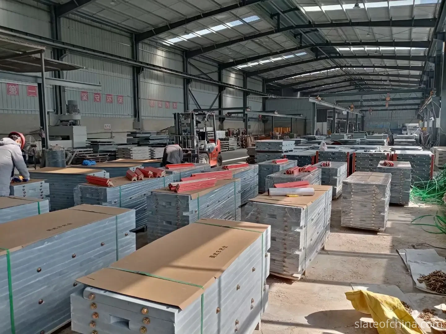 Packed billiard slate beds for 8-foot American pool tables stacked on wooden pallets with green strapping and stretch film in a factory warehouse, a red forklift and workers in the aisle