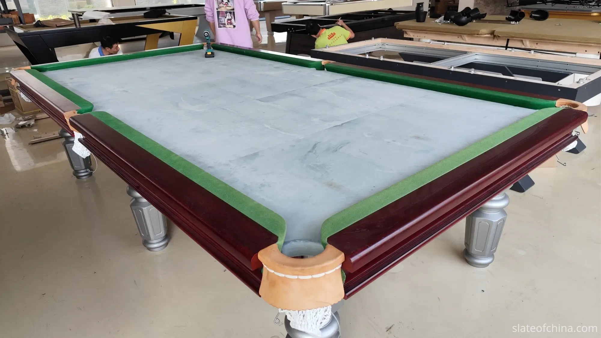 Three-piece billiard slate bed installed on a pool table during assembly in a workshop, seams visible between the bare grey panels, a cordless drill resting on the stone