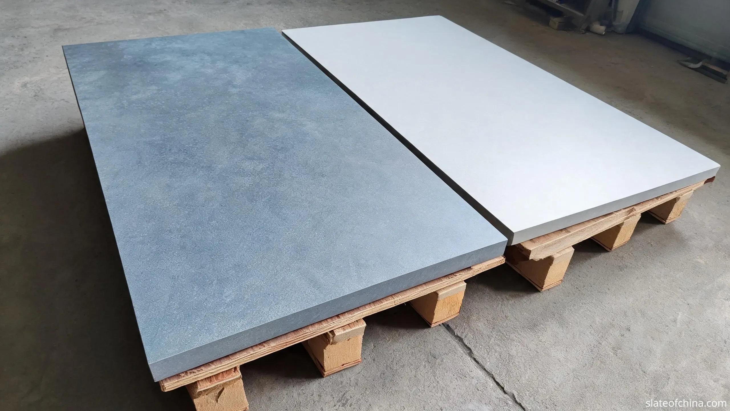 Cool blue-grey natural slate panel with subtle mottling and a light grey composite board lying side by side on wooden pallets in a workshop