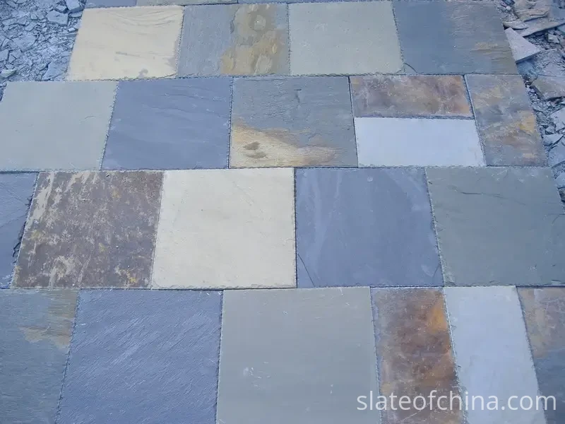 Paving Slate Sizes & Thicknesses 1 Mixed-format natural slate paving slabs laid out flat on quarry gravel in rows, square and rectangular cleft-face pieces in varied blue-grey and warm rust tones with natural split edges