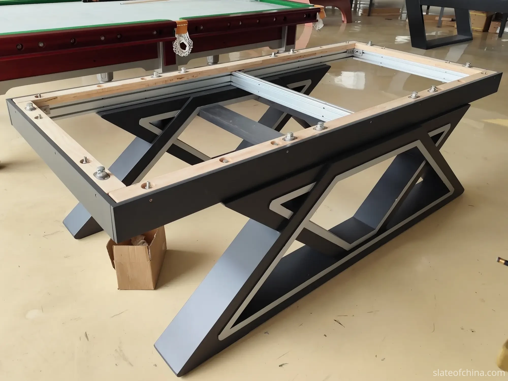 Empty billiard table frame prepared for slate installation in a workshop, mounting bolts in place on the support ledge, finished tables in the background