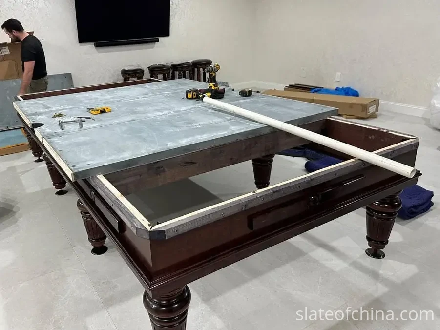 Long rigid straightedge laid across the seam between two bare grey billiard slate panels to check joint height alignment, installation tools resting on the bed