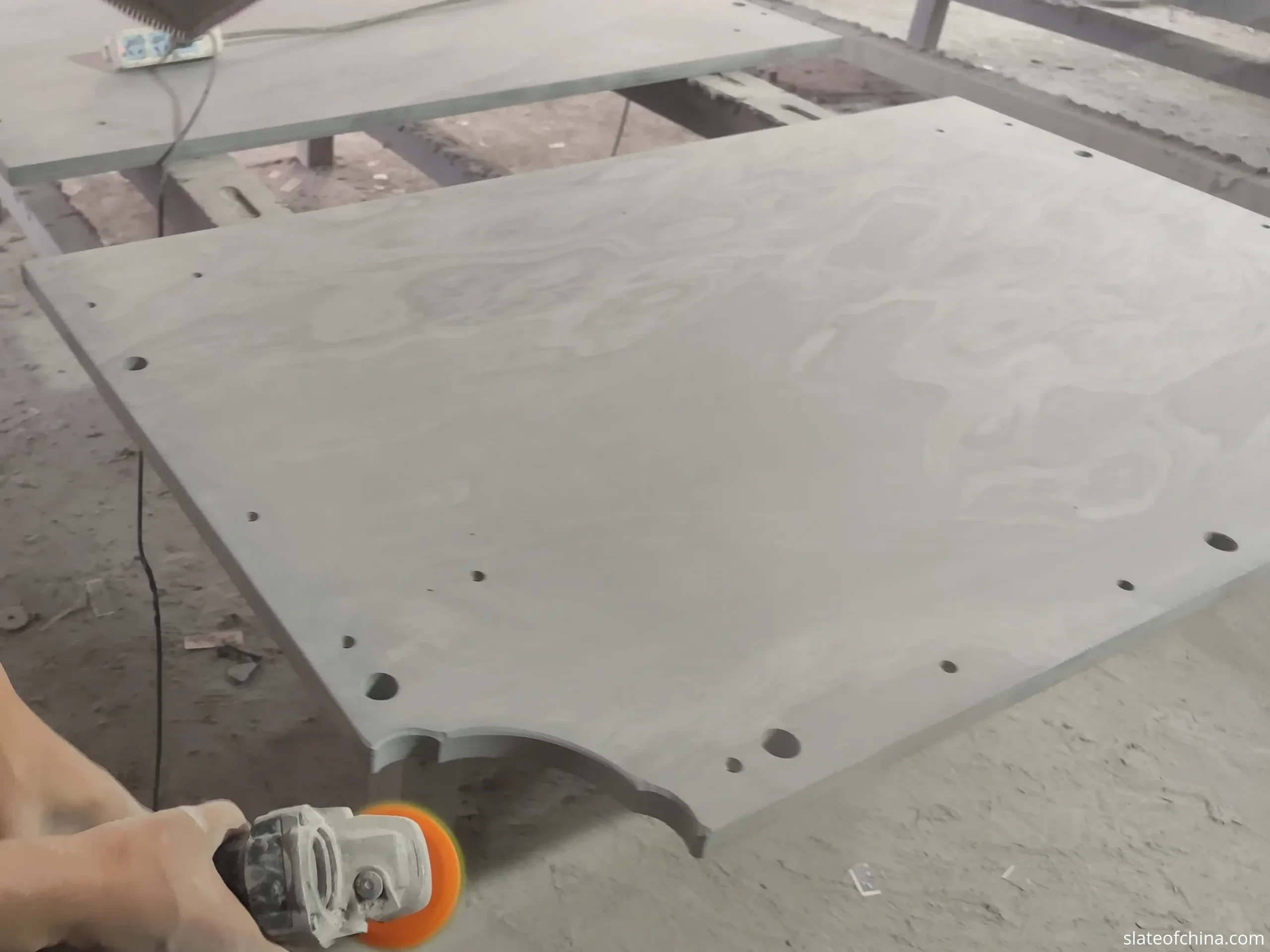 Professional tournament slate bed edge finishing on inspection bench Real factory photo of a worker with an angle grinder finishing the edge of a natural slate billiard bed panel on a heavy steel inspection bench, pre-drilled holes and pocket cutout visible