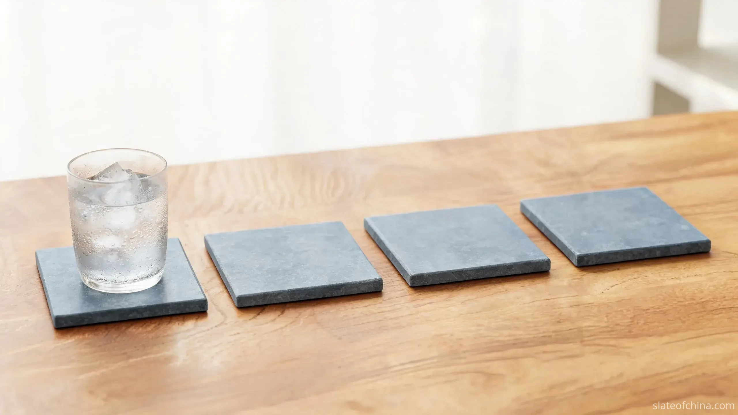 Slate Coasters from Jiujiang 1 Illustrative view of four square natural slate coasters with straight cut edges on an oak tabletop, a cold glass with condensation standing on the first