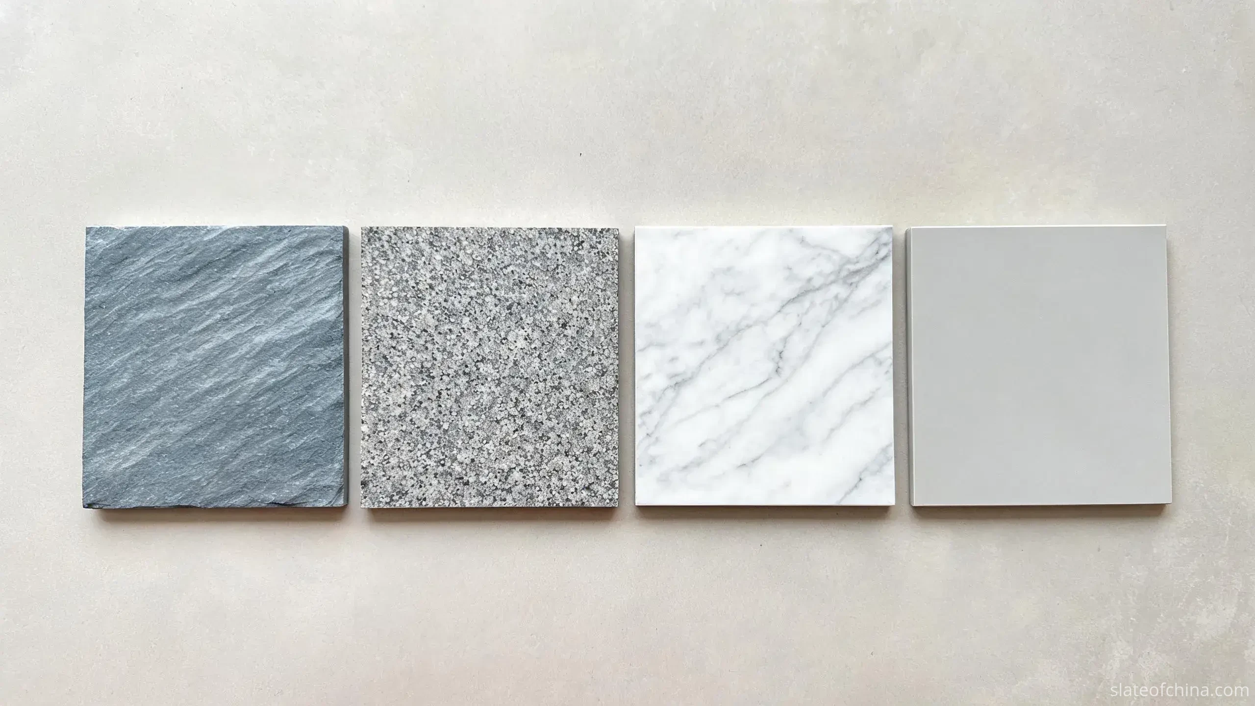 Slate Flooring vs Other Stone Flooring 1 Four square flooring samples side by side on a light worktable - blue-grey cleft natural slate, speckled grey granite, veined white marble and uniform matte porcelain tile