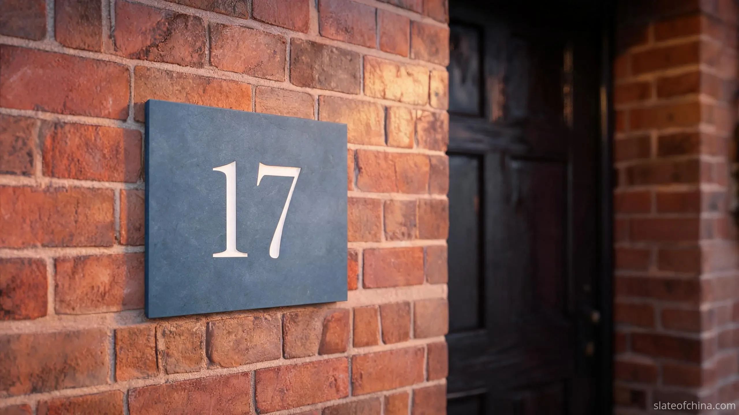 Slate Signs & House Number Plates 1 A dark blue-grey slate house number plate with pale engraved numerals fixed to a warm brick wall in soft dusk light