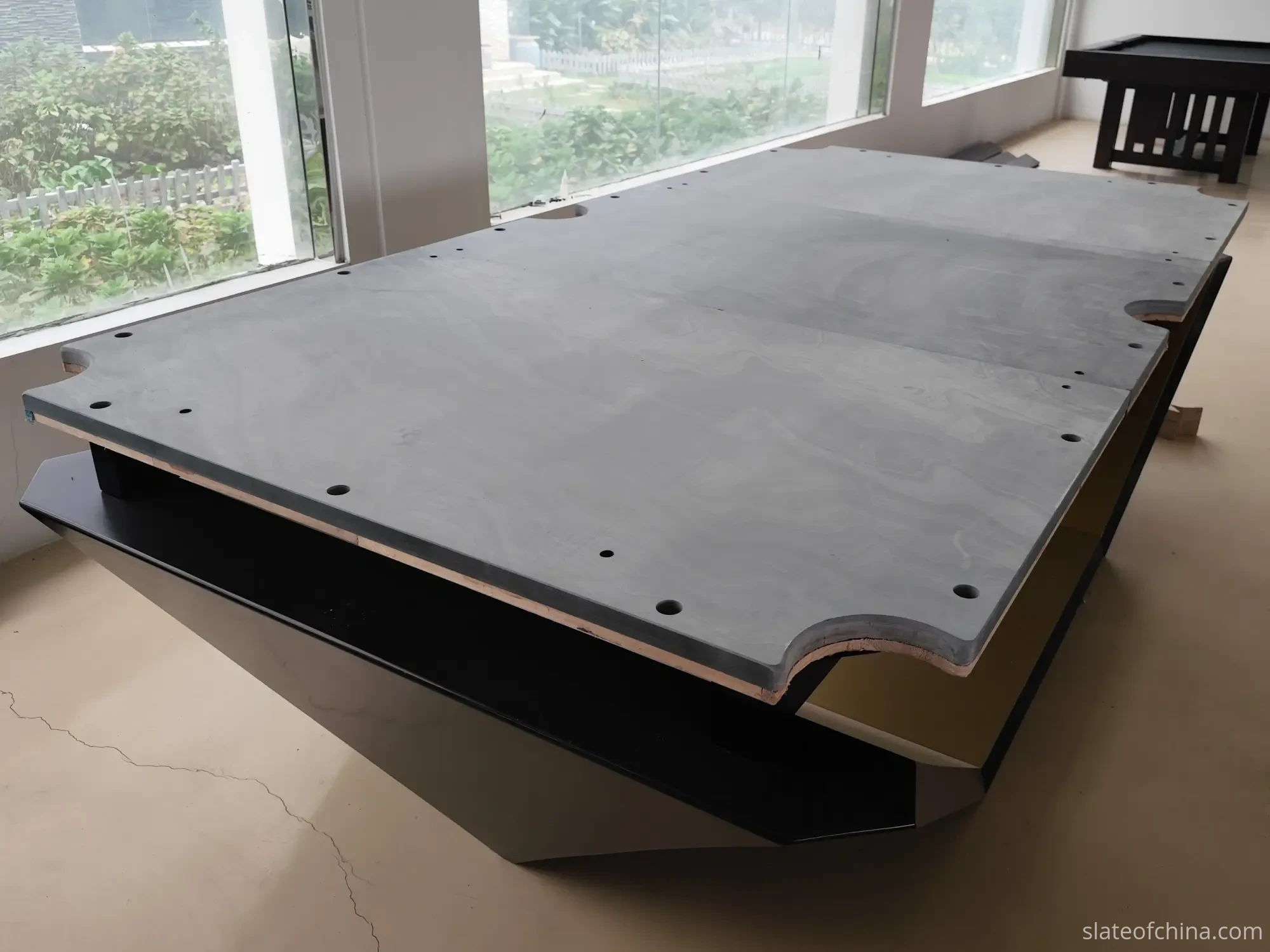 How Slate Flatness Affects Ball Roll 4 Three-piece honed billiard slate on a black table base under raking daylight from floor-to-ceiling windows, subtle surface shading and pocket cutouts visible